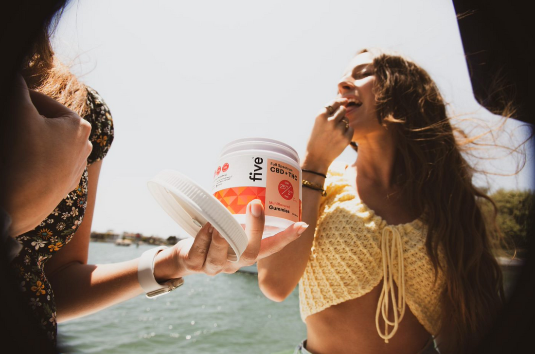 Person holding a container labeled 'five' with 'Full Spectrum CBD + THC' and 'Multiflavor Gummies', another person about to eat a gummy outdoors near water.