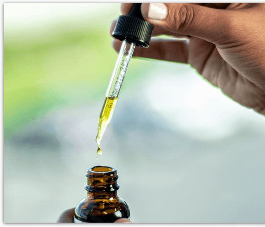 Hand holding a dropper with yellow liquid dropping into a brown glass bottle.