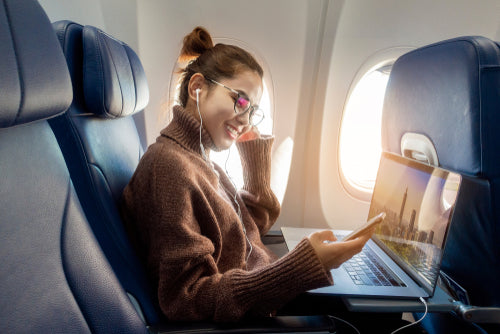 Woman wearing glasses and earphones, smiling while using a laptop on an airplane seat next to a window with sunlight.