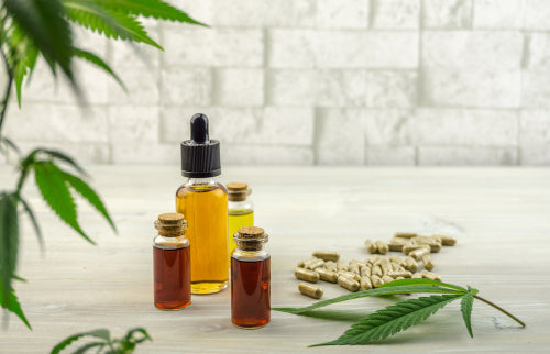 Three small bottles with amber and yellow liquids, cannabis leaves, and scattered capsules on a light wooden surface with a blurred brick wall background.