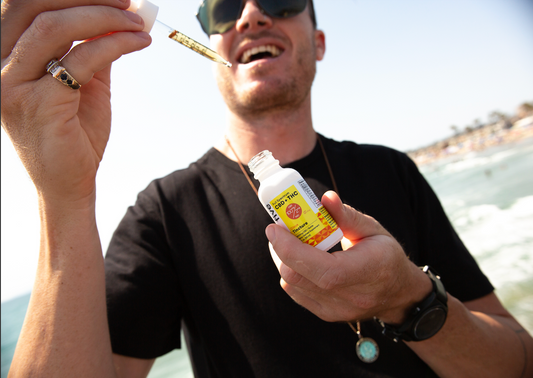 Man holding a CBD + THC tincture bottle labeled 'Full Spectrum CBD + THC 100% Natural Citrus Tincture' and a dropper near his mouth, wearing sunglasses and a black shirt at the beach.