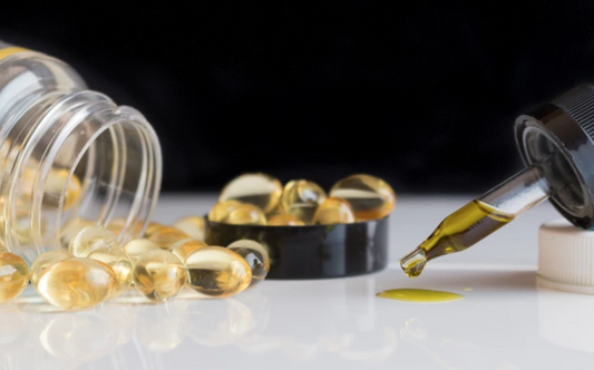 Capsules spilled from a clear bottle next to a dropper releasing a drop of yellow oil on a white surface with a black background.
