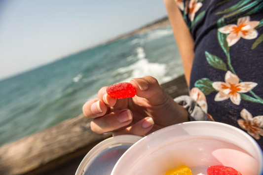 Hand holding a red gummy candy above a container with more red and yellow gummies, with a body of water and a person wearing a floral shirt in the background.