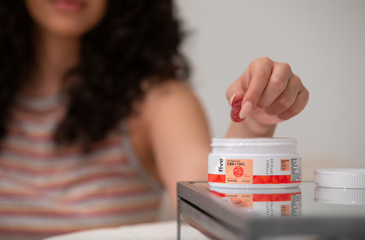 Hand holding a red gummy above a white container labeled 'five Full Spectrum CBD + THC Multi-flavored Gummies 25 mg CBD 2.5 mg THC per gummy 20ct Bottle' on a reflective surface