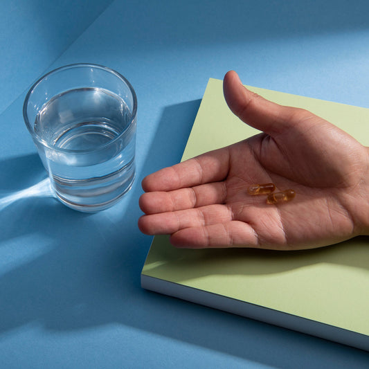 Hand holding two amber softgel capsules beside a glass of water on a pale green notepad