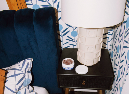 Bedside table with a white lamp, an open container of brown gummies, and a blue velvet headboard with patterned pillows in the background.