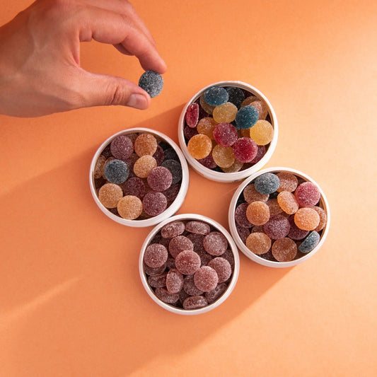 Hand holding a blue sugar-coated gummy candy above four white containers filled with assorted colorful sugar-coated gummy candies on an orange background