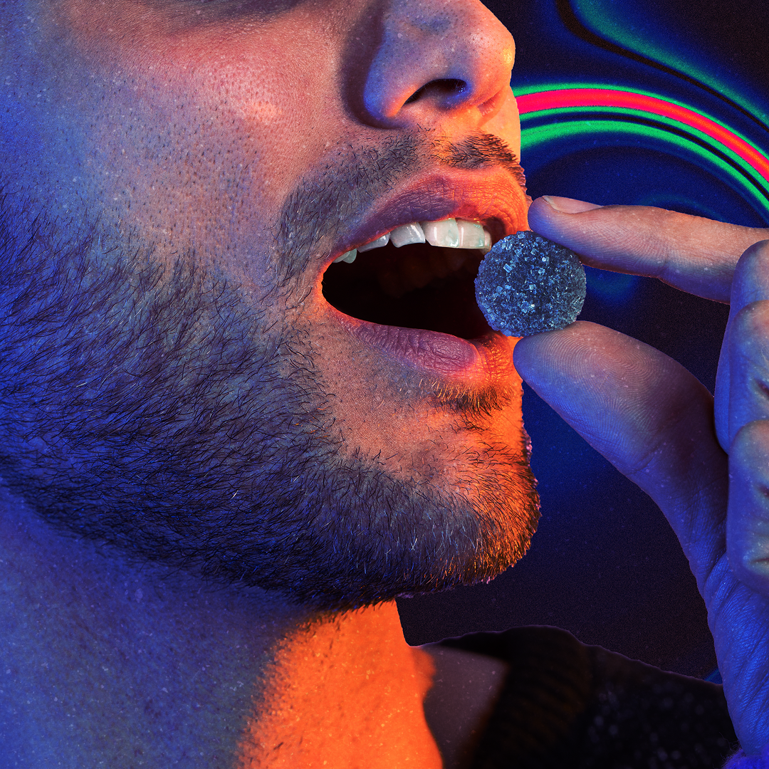 Close-up of a bearded man holding a sugar-coated cannabis edible near his open mouth against a neon-lit background.