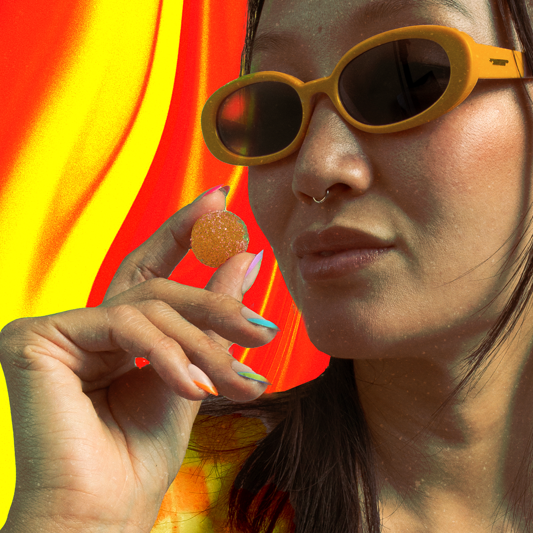 Close-up of a person in yellow sunglasses holding a sugar-coated gummy between fingers, red and yellow wavy background.