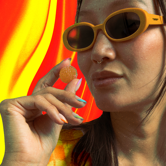 Close-up of a person in yellow sunglasses holding a sugar-coated gummy between fingers, red and yellow wavy background.