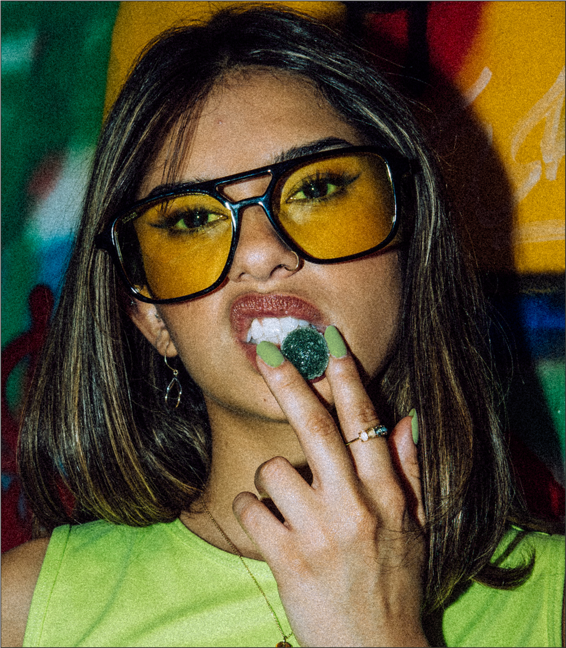Young woman wearing yellow-tinted glasses holding a green gummy near her mouth