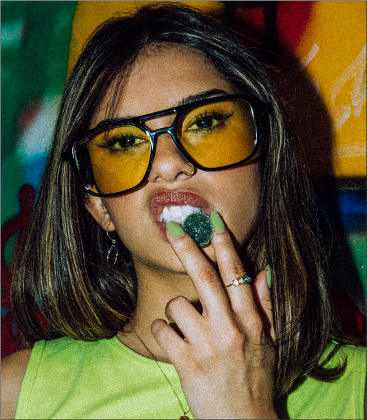Young woman wearing yellow-tinted glasses holding a green gummy near her mouth
